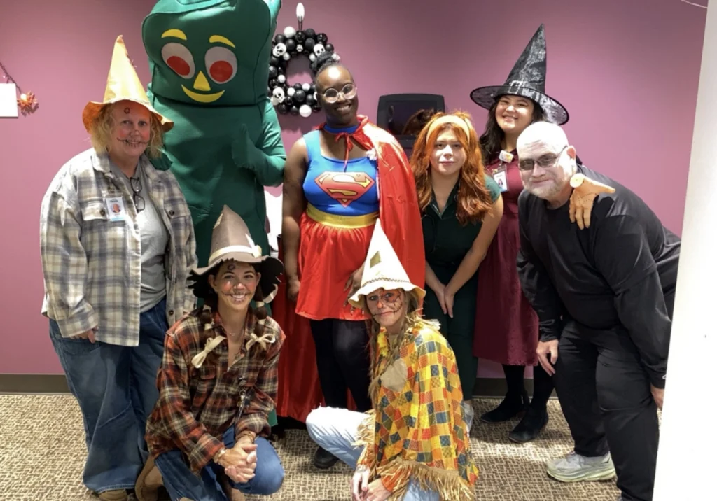 Eight people in colorful costumes, including Gumby, Supergirl, a witch, and several scarecrows, pose and smile together in front of a purple wall decorated with a black and white Halloween wreath.
