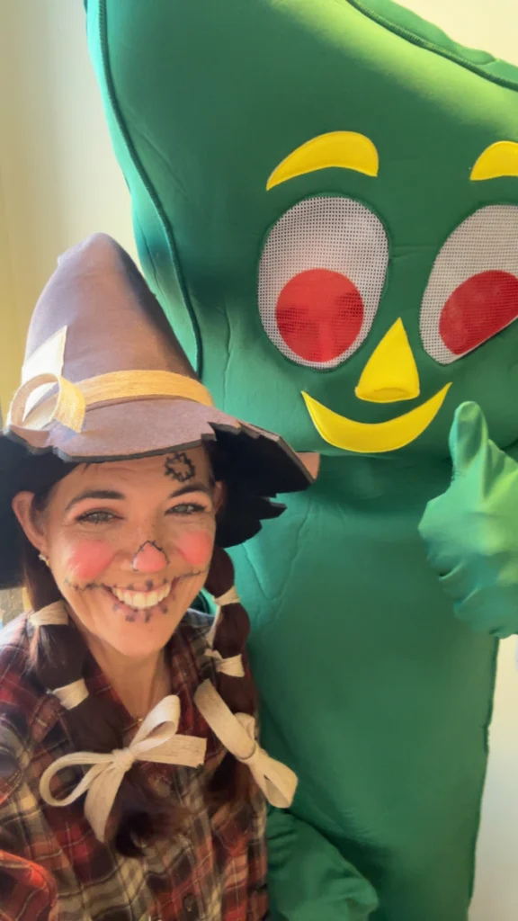 A person dressed as a scarecrow with face paint and a hat smiles next to someone in a large green Gumby costume, both posing happily for the camera indoors.