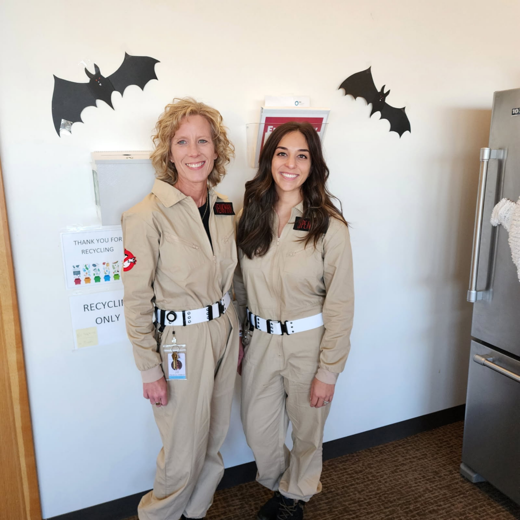 Two women dressed in beige Ghostbusters costumes stand side by side, smiling. Black paper bats are decorated on the wall behind them, and a recycling sign and a refrigerator are visible in the background.