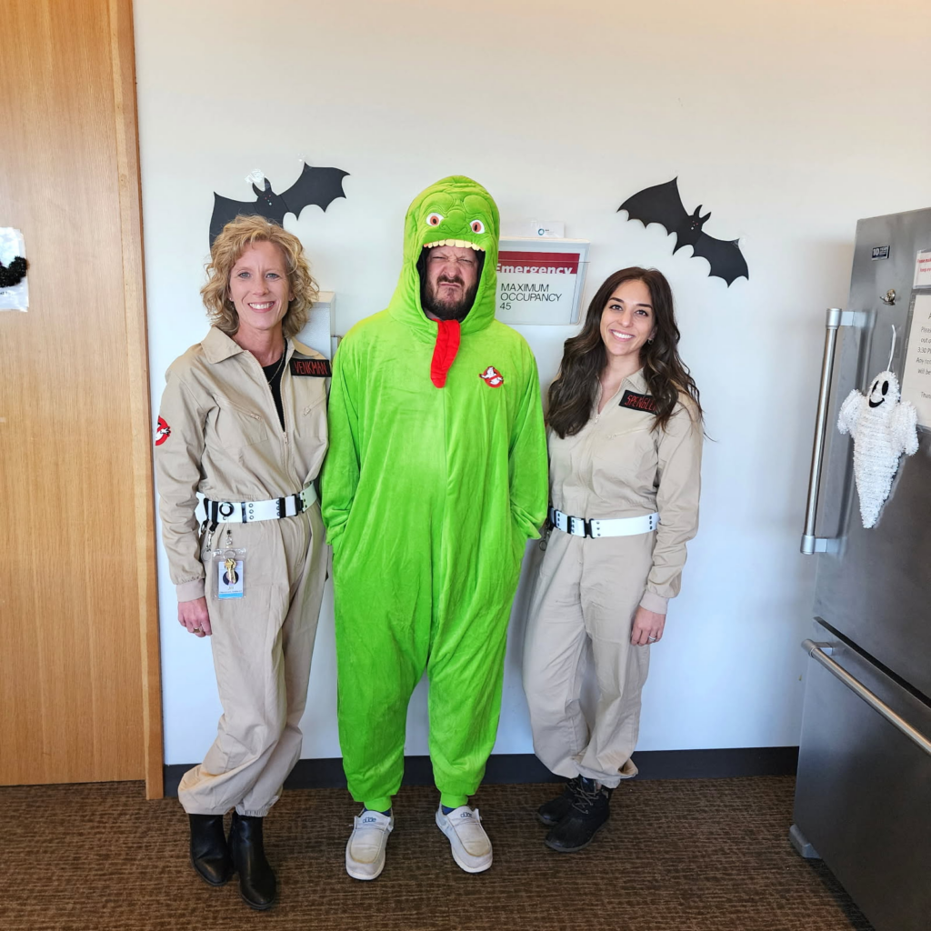 Three adults in costumes stand indoors. Two women wear Ghostbusters uniforms, while the man in the middle wears a bright green monster suit with a red tongue. Black bat decorations and a ghost are on the wall and fridge behind them.
