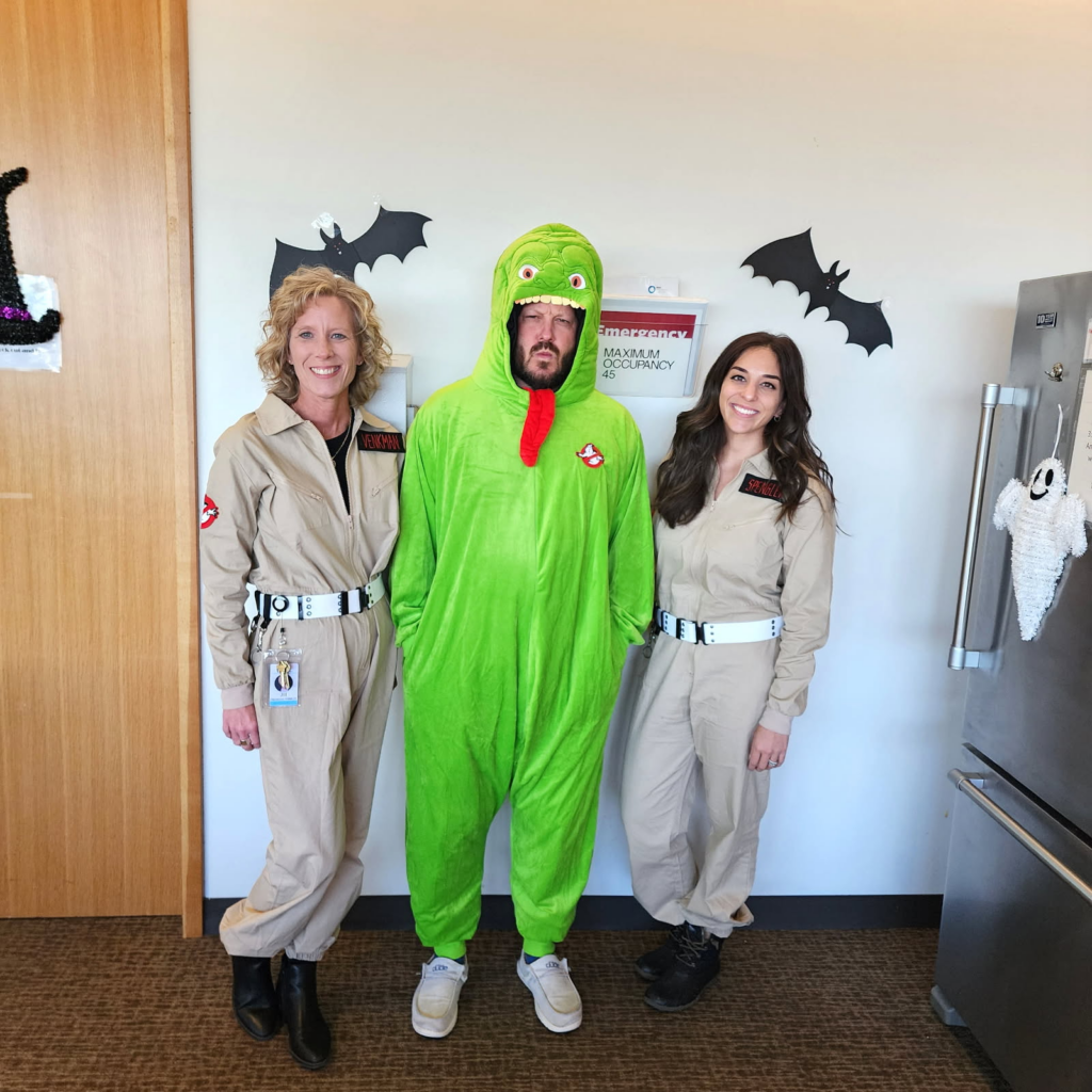 Three people stand in front of a wall decorated with black paper bats. Two women wear Ghostbusters costumes, and a man in the middle wears a bright green dinosaur costume with a red tongue hanging down.