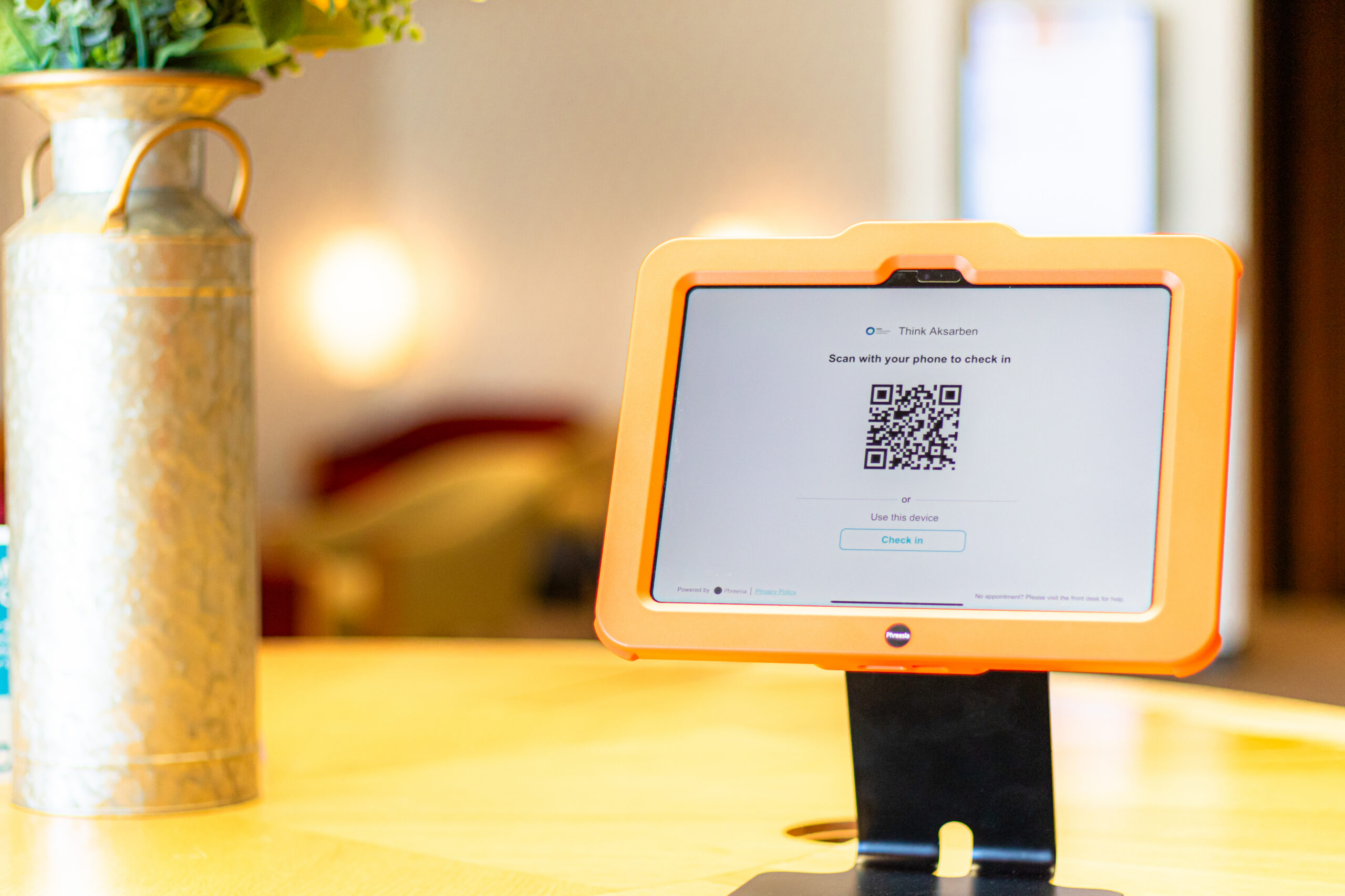 A tablet in an orange case displaying a QR code sits on a stand atop a round wooden table; a tall vase with yellow flowers is in the background.