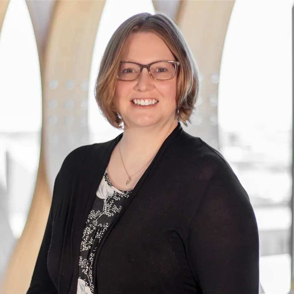 A smiling Alexandra Harvey, LIMHP, with short light brown hair and glasses, wearing a black cardigan over a patterned top, stands in front of a bright background with curved wooden beams.