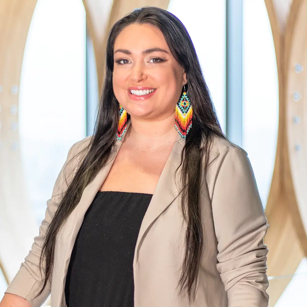 Alison Kampe, LIMHP, with long dark hair, wearing a tan blazer over a black top and colorful beaded earrings, smiles while standing in front of a bright window with a modern wooden design in the background.