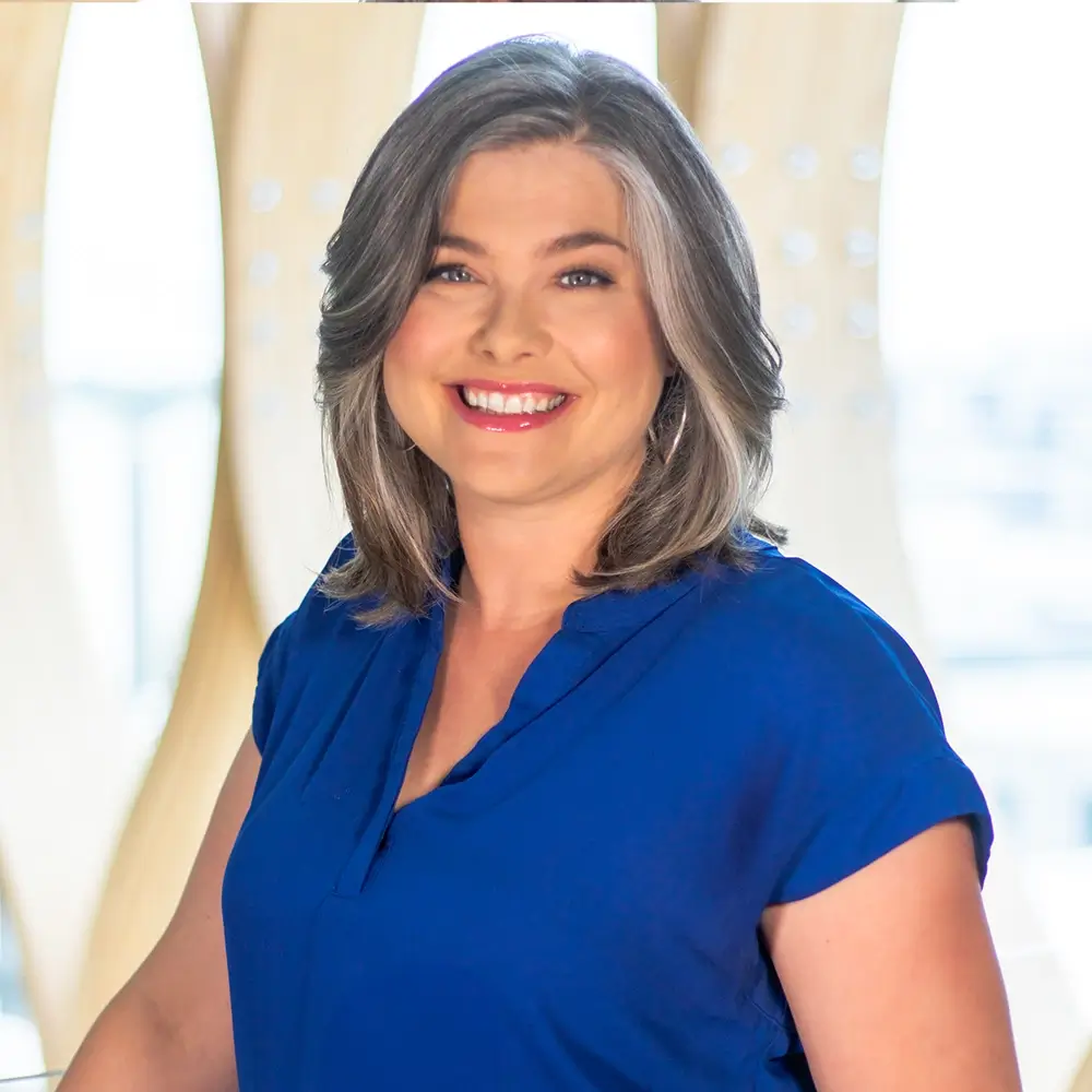 Erica Irwin, LIMHP, with shoulder-length gray hair, smiles at the camera. She is wearing a bright blue, short-sleeve blouse and stands in front of a softly lit, modern background with light wooden panels.
