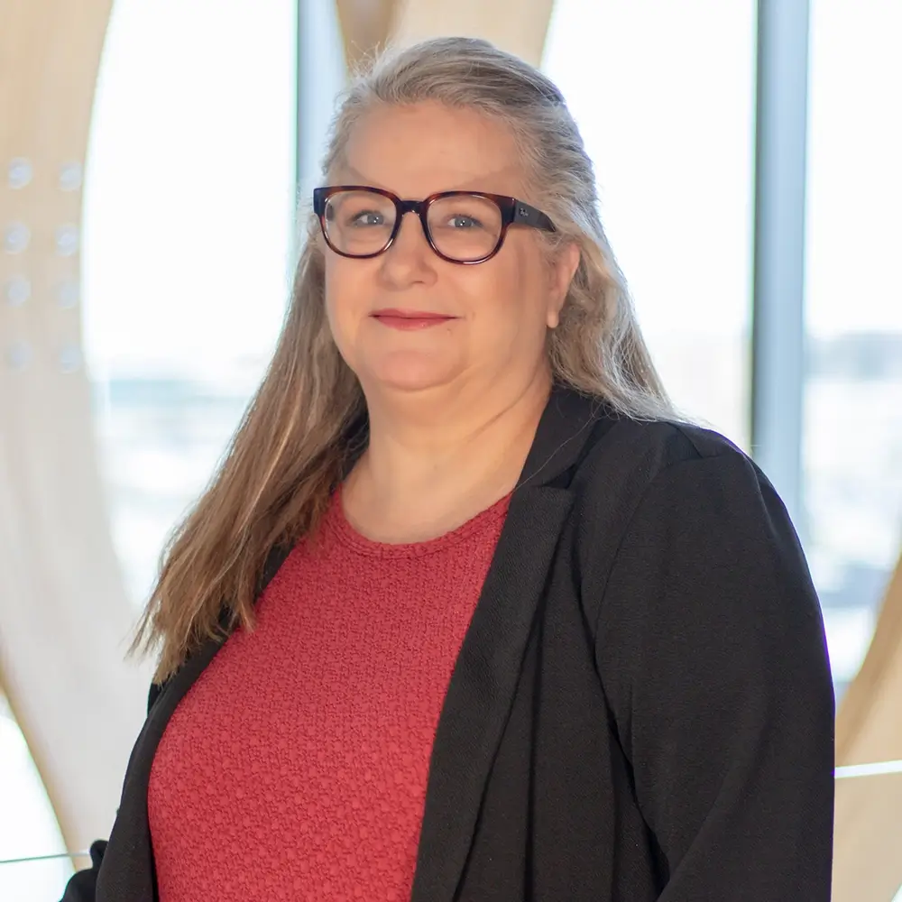 Holly Eckhart, LIMHP, a woman with long gray hair and glasses, wears a black blazer over a red textured top as she stands indoors with large windows and natural light in the background.