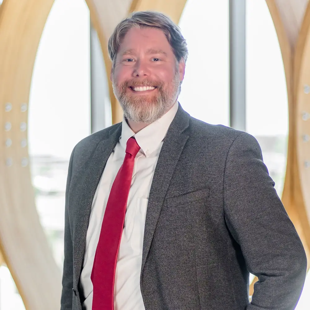 Jameson Bock, LIMHP, a smiling man with a beard, wears a gray suit, white shirt, and red tie as he stands before tall windows with curved wooden frames and natural light in the background.