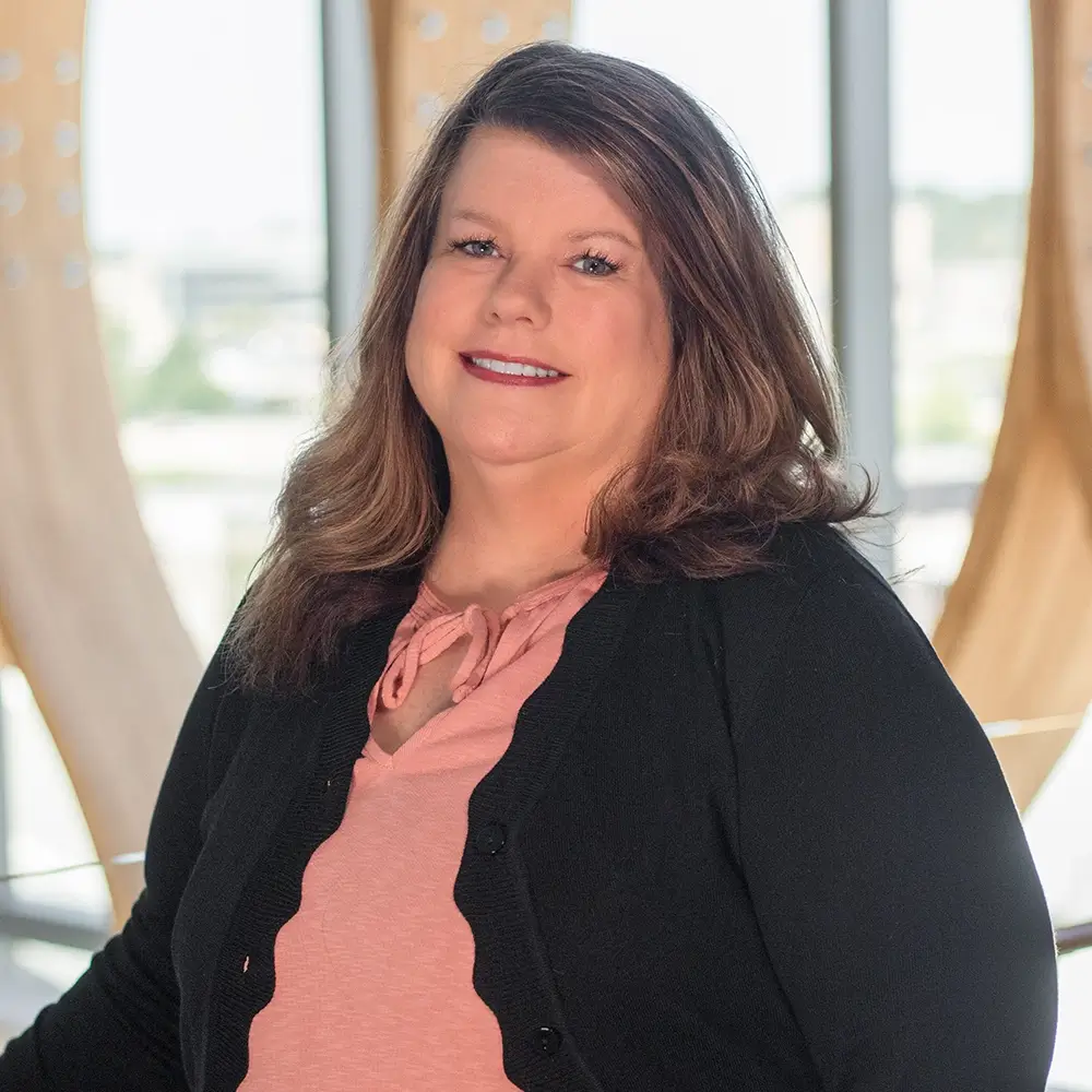 Jodi Ramold, LIMHP, a woman with long brown hair, smiles in a pink top and black cardigan while standing indoors in front of large windows with modern wooden architectural details.