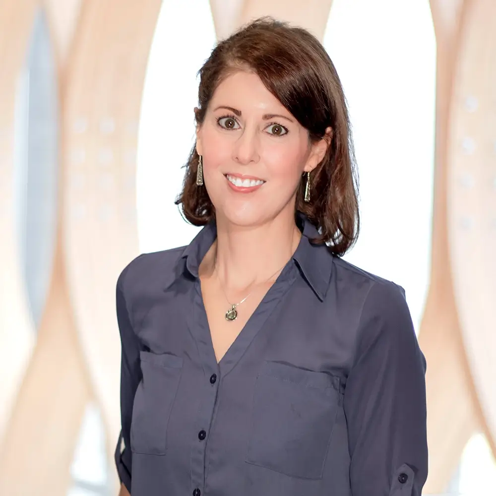 Lyndsay Dean, DNP, with shoulder-length brown hair, wearing a navy blue blouse and earrings, stands smiling in front of a light-colored, abstract patterned background.