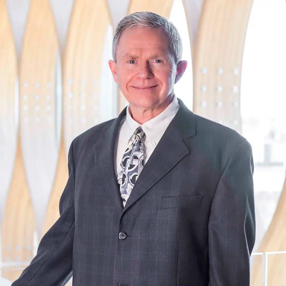 A smiling older man with short gray hair, Rodney L. Nitcher, DO, is wearing a dark suit, white shirt, and patterned tie, standing indoors in front of a modern background with wooden arches.
