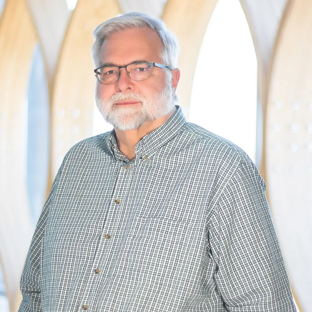 Russell Rosenquist, LICSW, a man with gray hair, a white beard, and glasses wearing a green and white checkered shirt stands in front of a bright background with large, arched window-like structures.