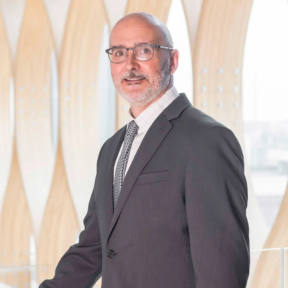 Brian Poole, MD, a man with glasses and a beard, dressed in a dark suit, white shirt, and patterned tie, stands indoors in front of a modern, light-colored architectural background with curved wooden panels.