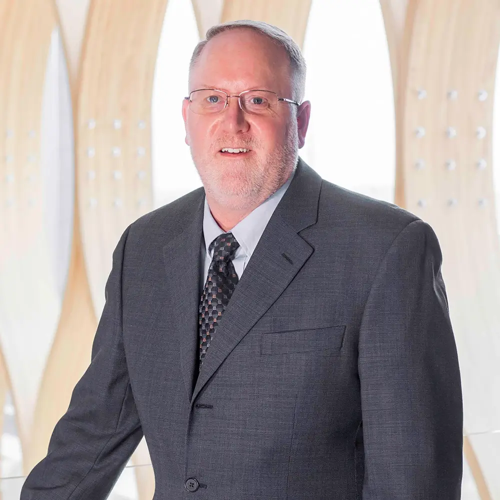 Brian Stoneking, APRN, a middle-aged man with short gray hair, glasses, and a trimmed beard, wears a gray suit, patterned tie, and light shirt while standing before a modern, light-colored abstract background.