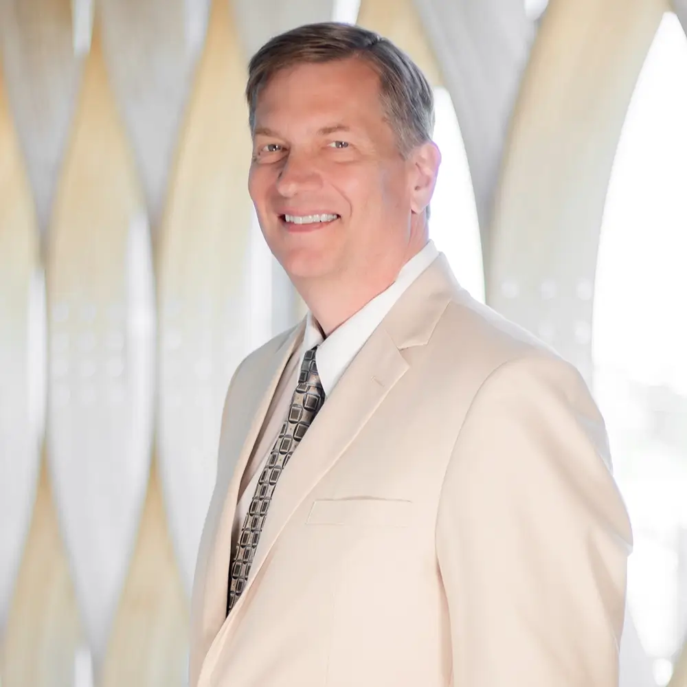 Dr. Charles Stoner, MD, wearing a light beige suit and patterned tie, smiles while standing indoors in front of a softly lit, modern backdrop featuring curved, vertical shapes.