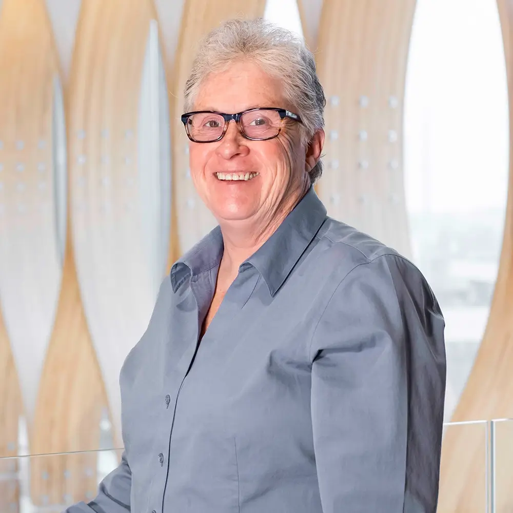 Cheryl Lindly, PA-C, an older woman with short gray hair and glasses, smiles warmly in a light blue button-up shirt while standing indoors against a modern, light-colored decorative background.