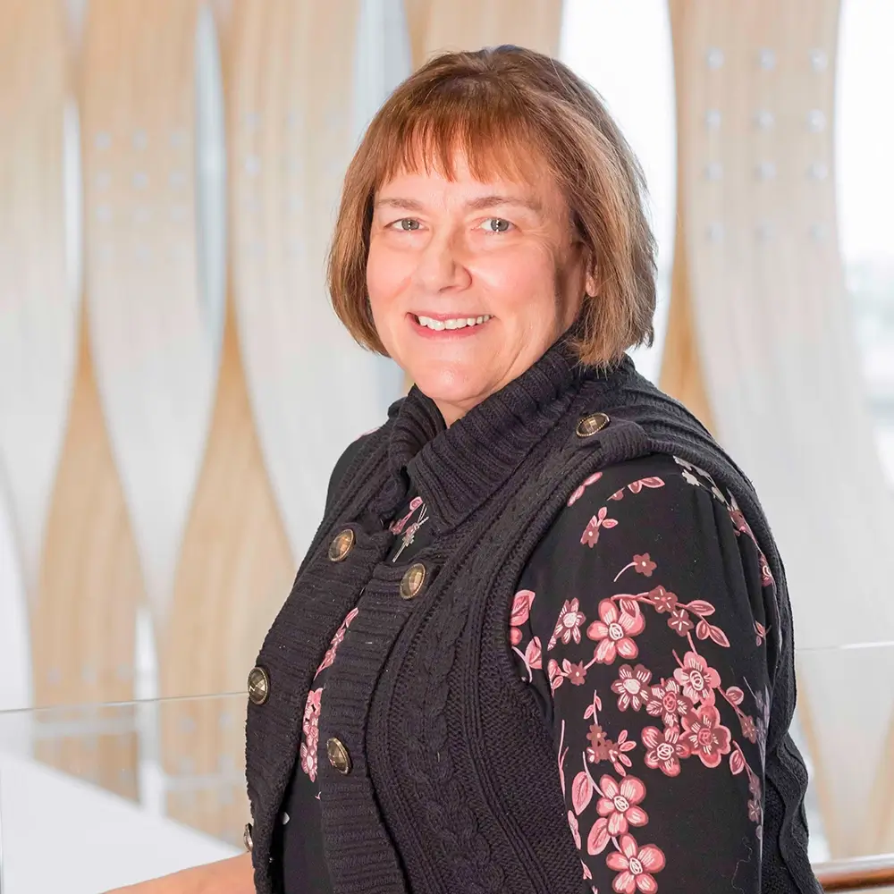 A woman with short brown hair, identified as Chris Jeffrey, MD, wearing a black sweater vest over a floral blouse, smiles while posing indoors against a background of curved light wood panels.