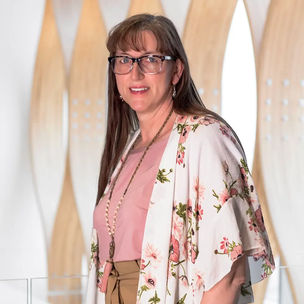 Christine Hunter, PA-C, with long brown hair and glasses, smiles at the camera. She wears a floral kimono over a pink top and tan pants, standing in front of a modern backdrop with curved wooden panels.