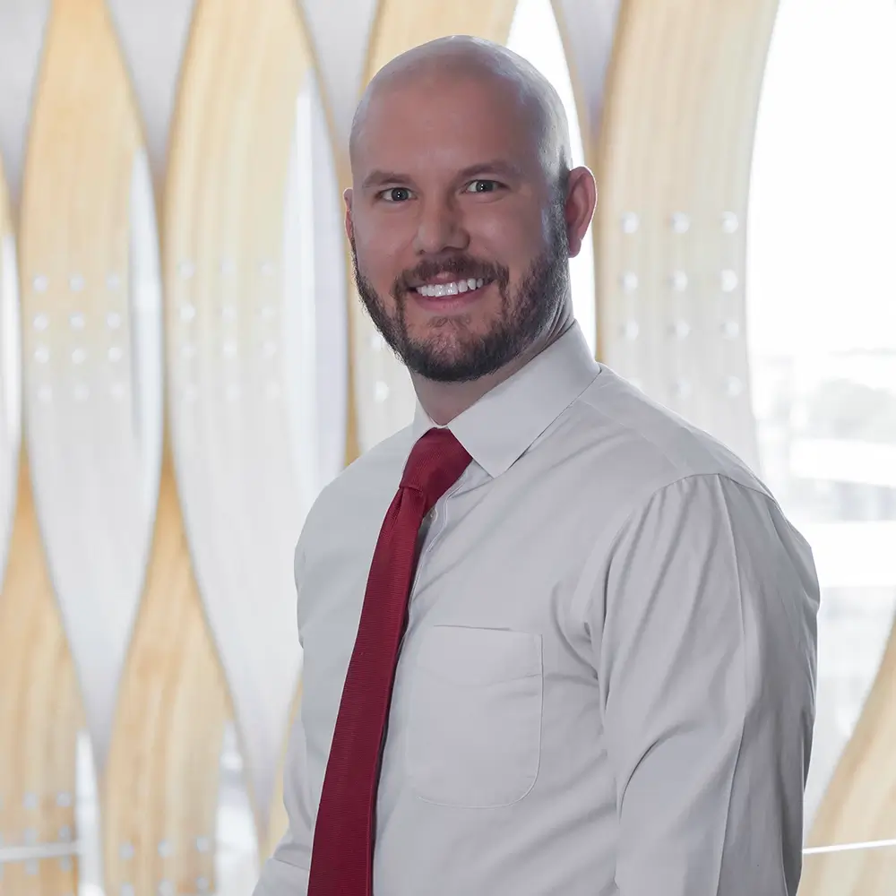 A man with a bald head and beard, identified as Austin Dudzinski, PharmD, smiles while standing in front of a modern, light-filled background with wooden panel accents, wearing a white dress shirt and red tie.