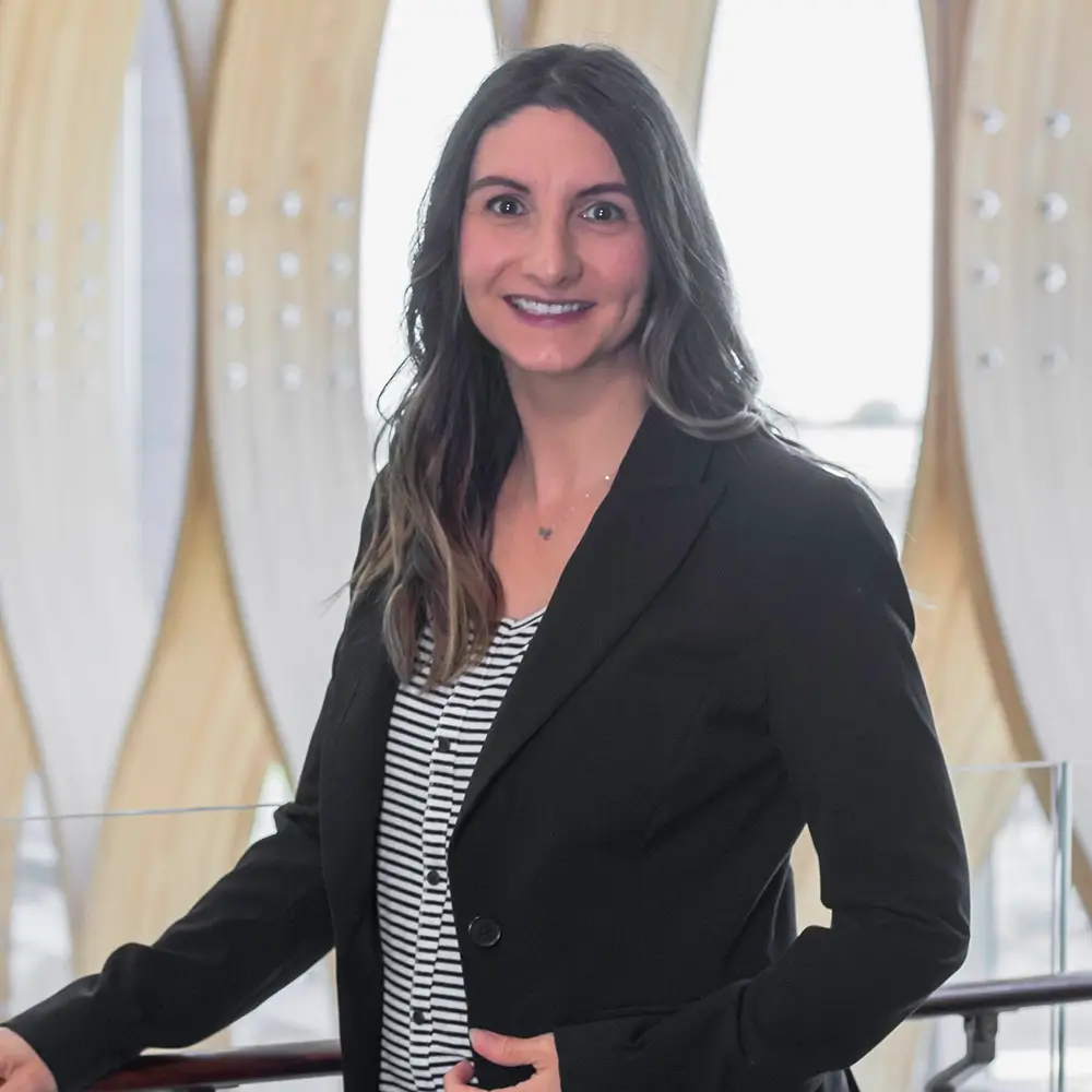 Kala Ray, PharmD, with long brown hair, wears a black blazer over a striped shirt as she stands indoors smiling at the camera. Tall, curved windows in the background let in soft natural light.