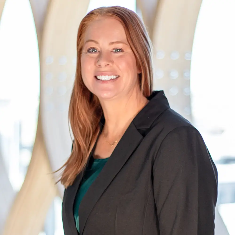 Maggie Hitzeman, PharmD, with long red hair, is smiling in a black blazer over a dark green top, standing in front of a modern, light-colored abstract background.