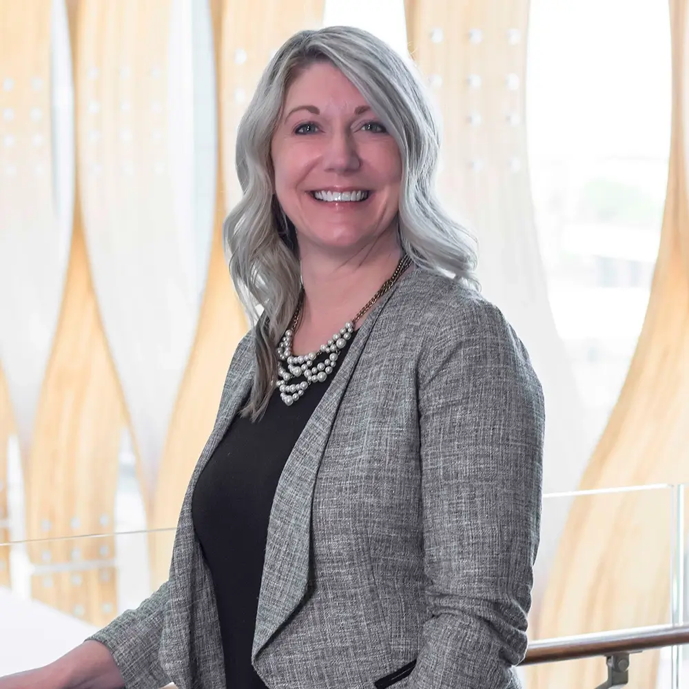 Mandi Russell, PharmD, a smiling woman with long, wavy blonde hair, wears a gray blazer, black top, and pearl necklace as she stands indoors before a modern backdrop of vertical wooden panels and soft natural light.