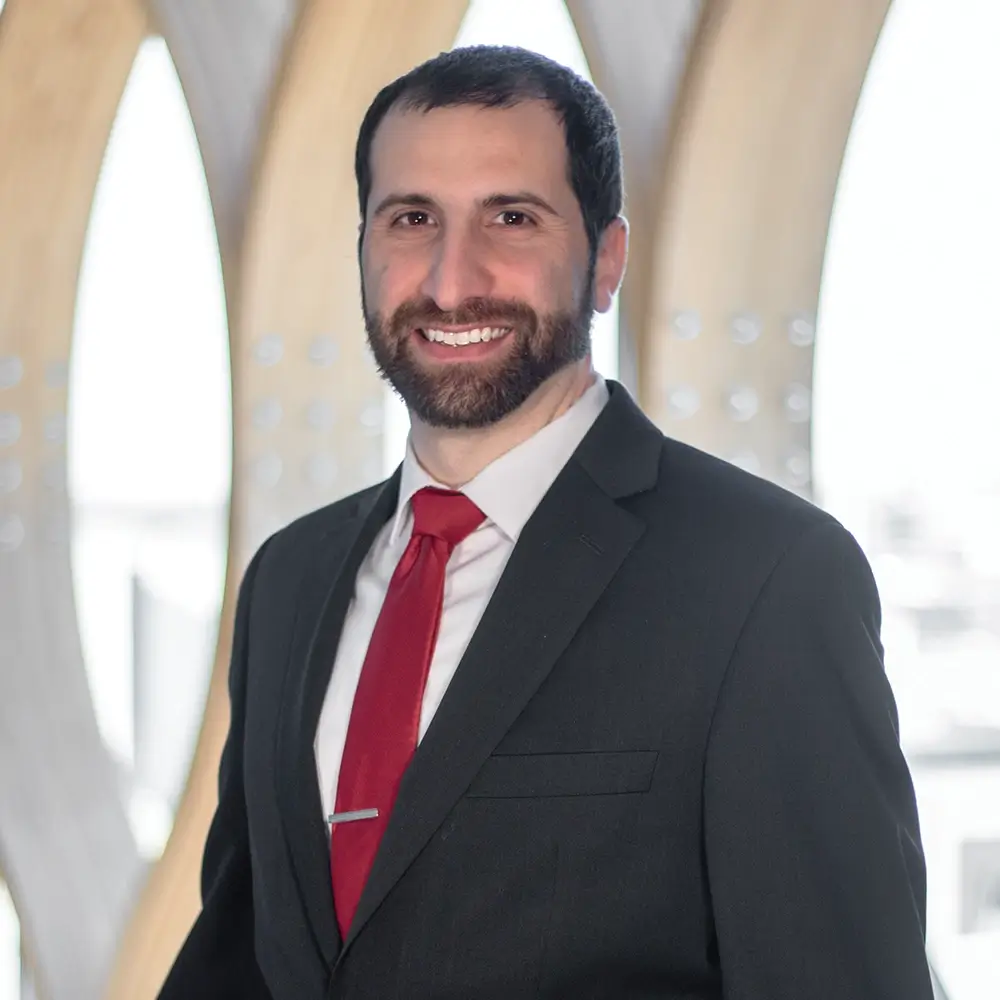 Mark Farrage, PharmD, a man with dark hair and a beard, wearing a black suit, white shirt, and red tie, stands smiling in front of a light, modern architectural background with curved wooden elements.