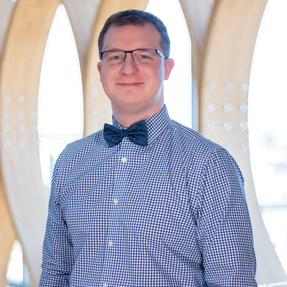 Michael Popkov, PharmD, wearing glasses, a blue and white checkered shirt, and a navy bow tie, stands smiling in front of a modern, light-filled background with curved wooden features.