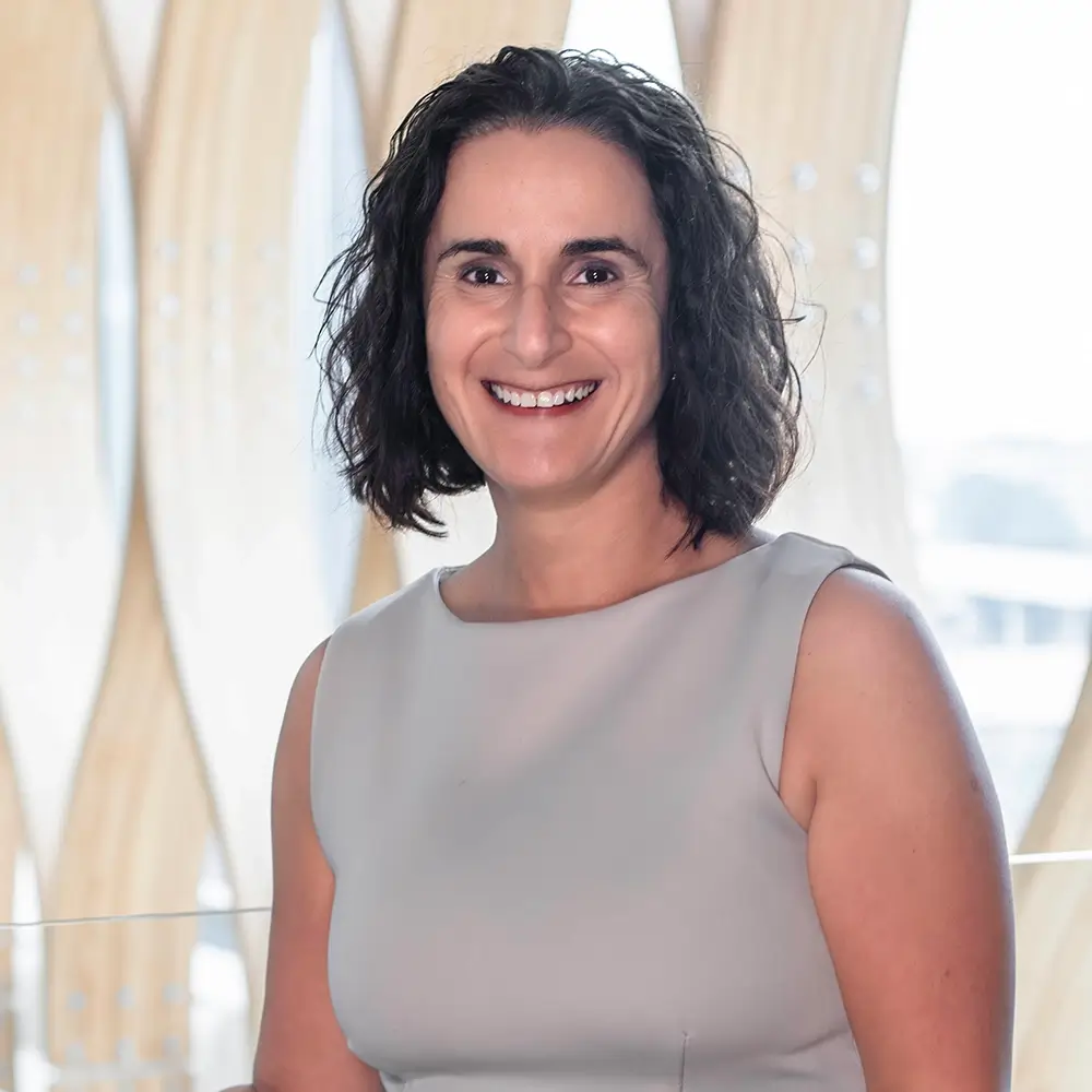 A woman with curly dark hair, wearing a sleeveless light gray top, smiles at the camera. Nairi Kasparian, PharmD, stands indoors in front of a wavy, light-colored patterned background.