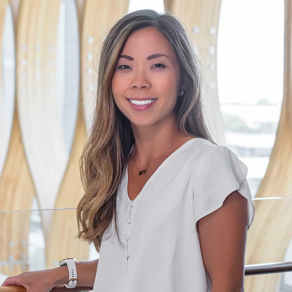 Shelley Lippold, PharmD, with long brown hair, wearing a white short-sleeve blouse and a watch, smiles as she stands indoors in front of a modern, wavy architectural background bathed in natural light.