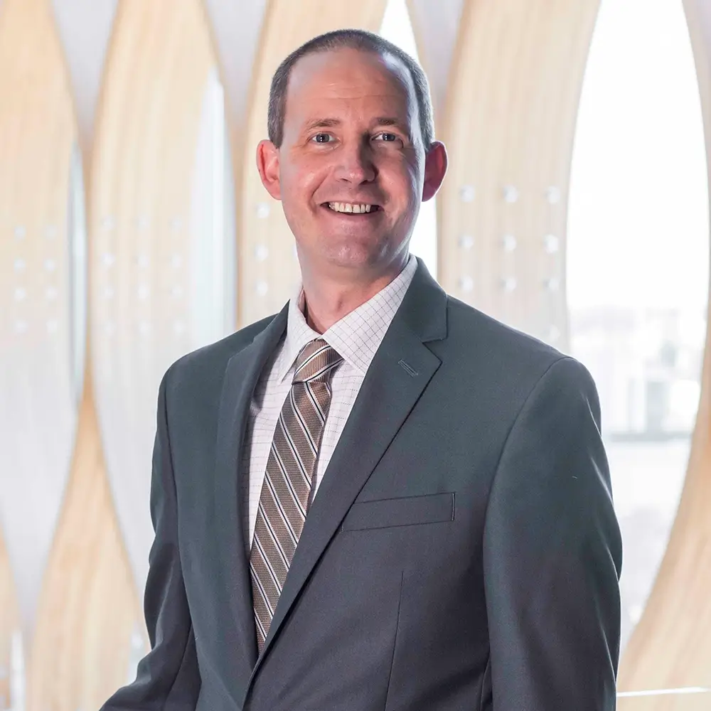David Thompson, MD, in a gray suit and striped tie stands smiling in front of a modern background with wooden arches and large windows.