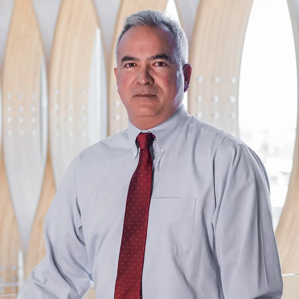 Dr. Doug Russell, MD, a middle-aged man with short gray hair, stands indoors in front of a modern wooden architectural background, wearing a light blue dress shirt and a red polka-dot tie.