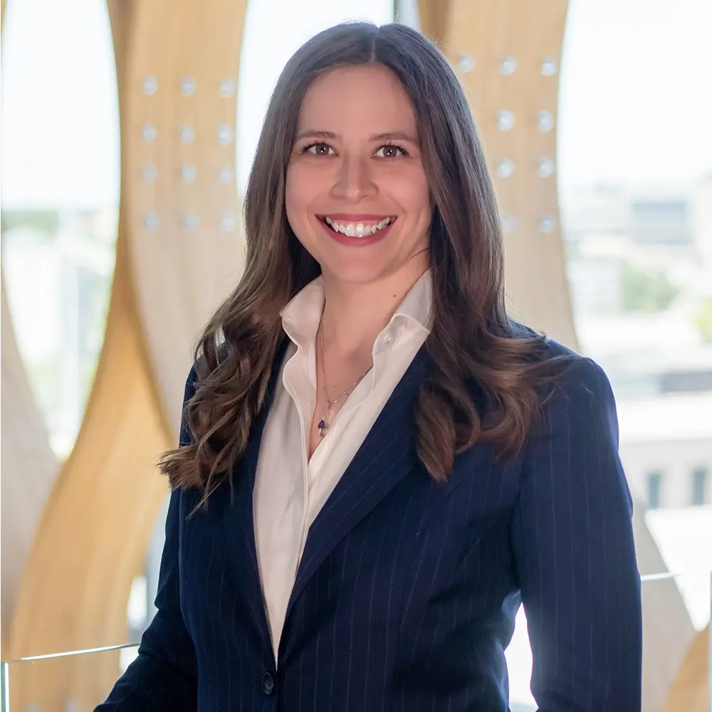 Dr. Erika Rothgeb, MD, with long brown hair, smiles warmly in a navy pinstripe blazer and white shirt while standing indoors before large wooden structures and windows offering a city view.