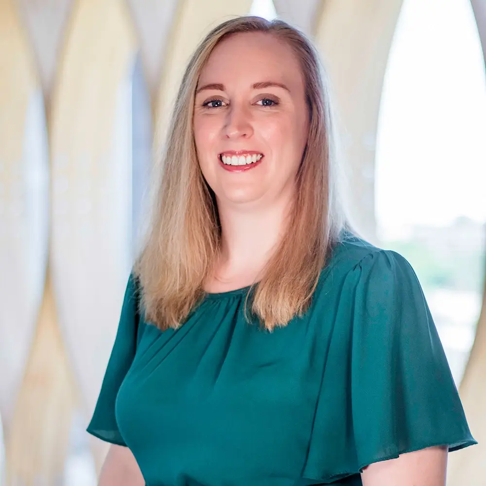 Erin Ray, DO, a woman with straight, shoulder-length blonde hair wearing a green blouse, smiles while standing indoors in front of a bright background with large, curved window panels.