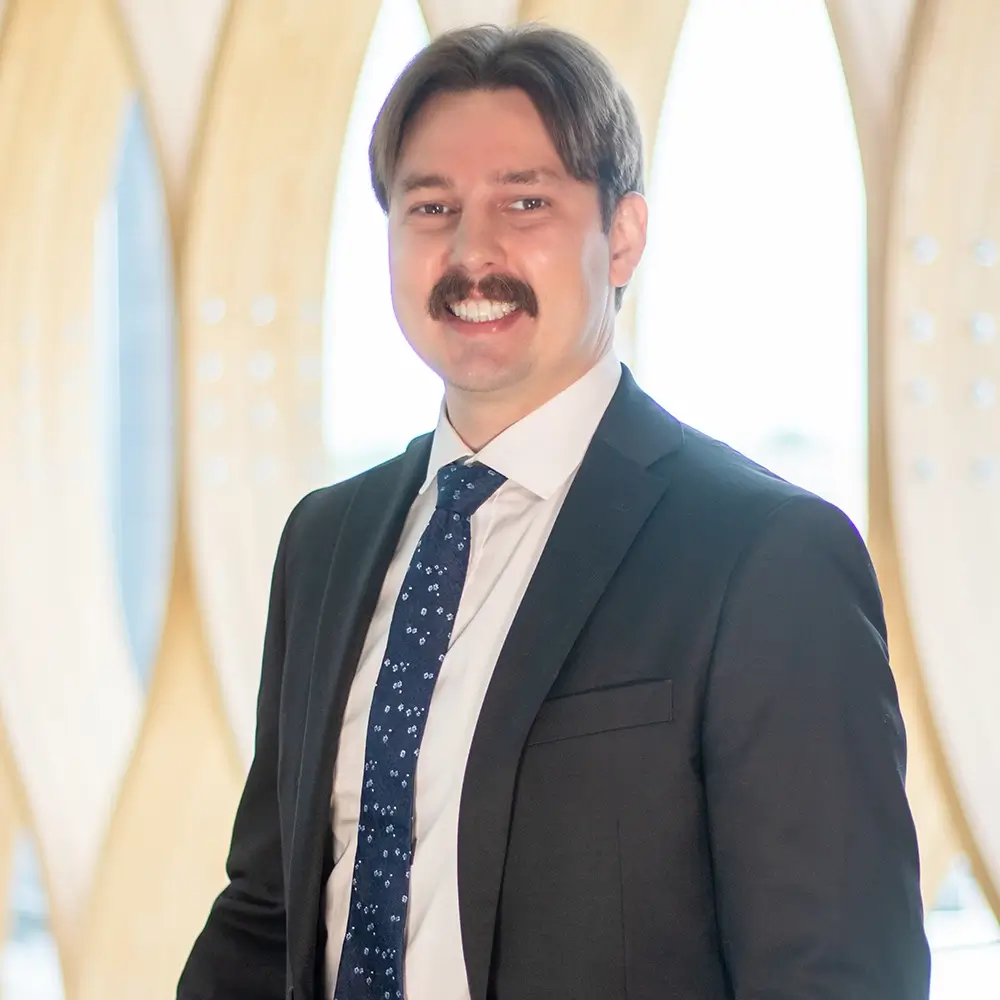 A man with dark hair and a mustache, Hleb Davydzenkau, MD, wears a black suit, white shirt, and patterned blue tie as he smiles in front of a modern, light-colored background featuring wavy wooden panels.