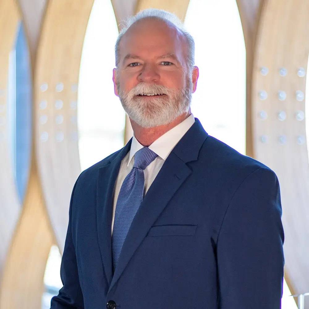 James Sullivan, MD, a man with white hair and a beard, stands in front of a modern, light-colored architectural background wearing a blue suit, white shirt, and blue tie.