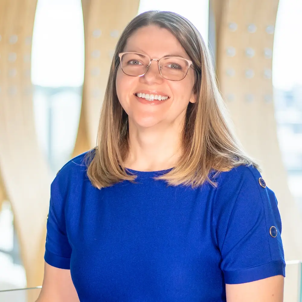 Dr. Jamie Vitamvas, MD, with straight light brown hair, wearing glasses and a royal blue top, smiles while seated indoors with a blurred wooden and glass background.