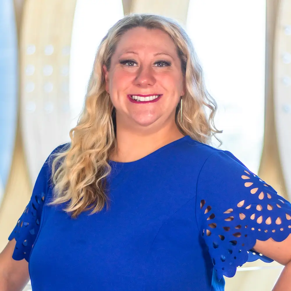 Dr. Jacqueline Hanson, MD, with long blonde hair and a bright blue dress with cut-out sleeves, smiles confidently at the camera against a softly blurred background with light and curved shapes.