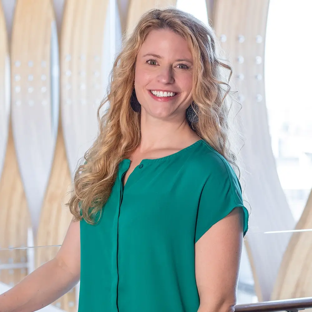 Jessica Grosse, PA-C, with long, wavy blonde hair and a teal blouse, smiles while standing indoors in front of a modern, decorative wooden wall.