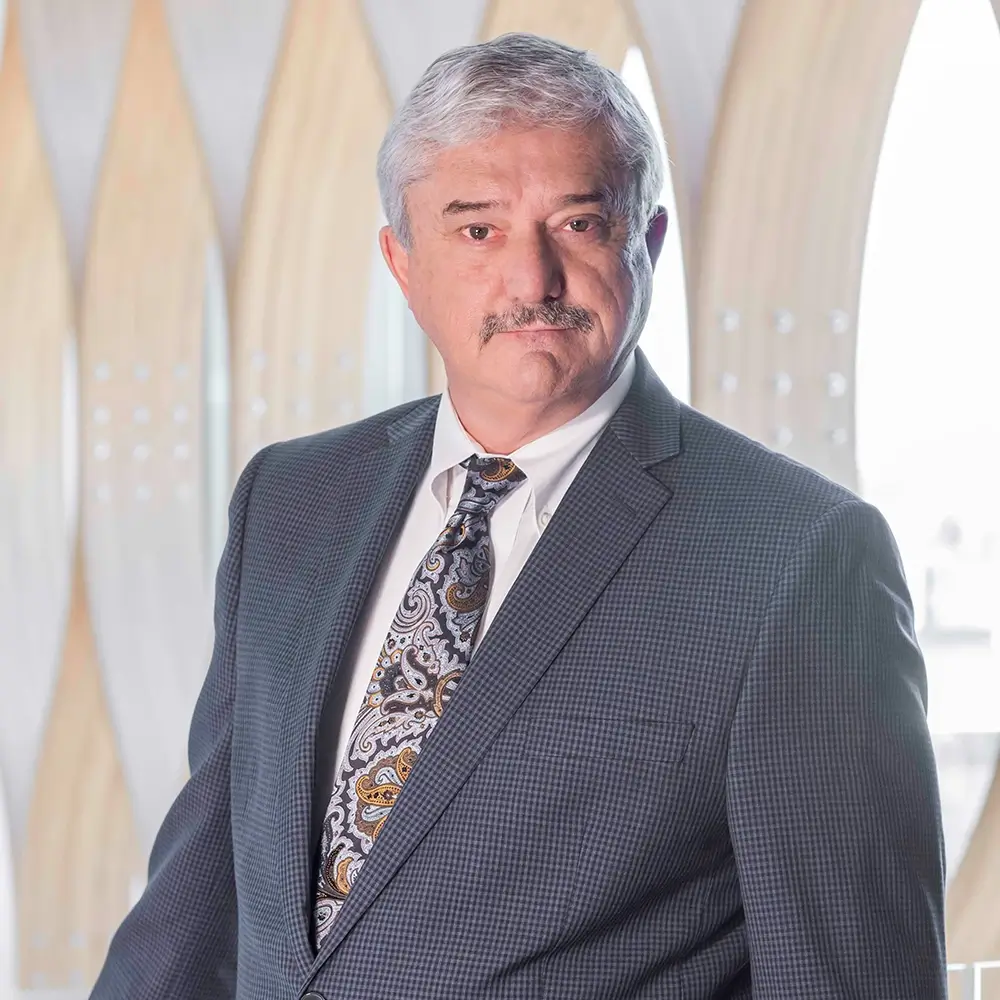 Dr. Joedy Istas, MD, an older man with gray hair and a mustache, stands indoors in front of a modern wooden and glass window, dressed in a checked suit, white shirt, and patterned tie.