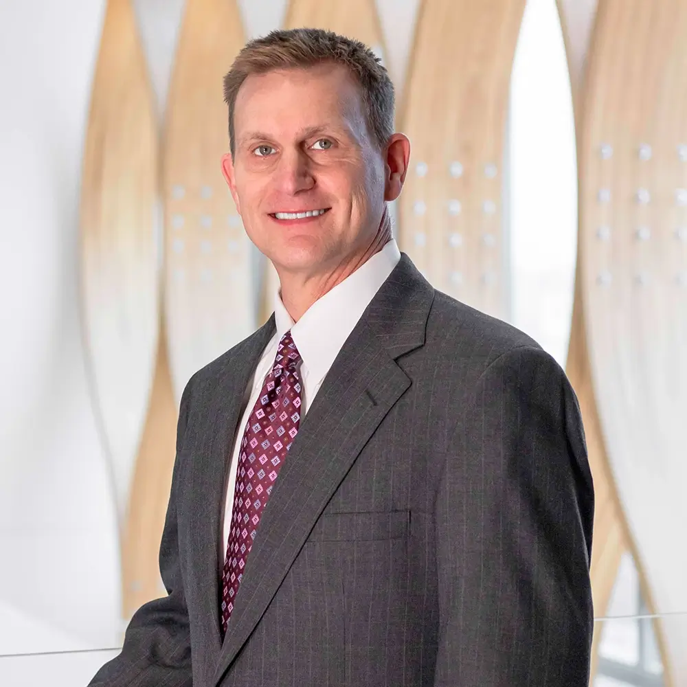 John Lewis, PA-C, in a gray suit with a patterned tie, stands indoors, smiling at the camera. Light wood panels and white accents create a warm, blurred background.
