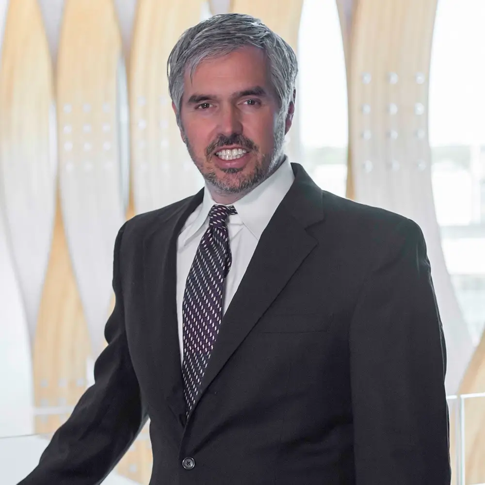 A man in a dark suit and striped tie stands indoors, smiling at the camera. Behind Joseph Shehan, MD, is a modern background with tall, curved wooden panels and soft natural light.
