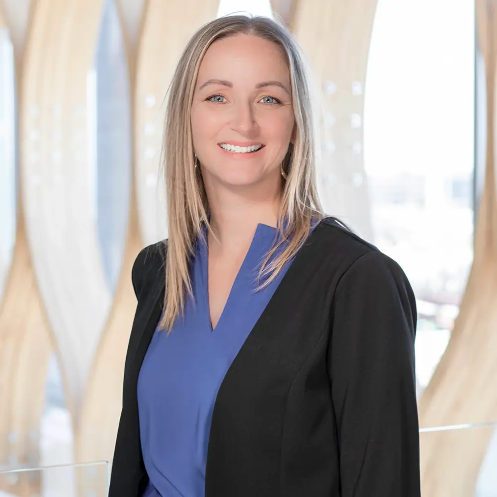 Katie Peterson, PA-C, with straight blonde hair, smiles in a blue blouse and black blazer while standing indoors against a modern light wood and glass background.