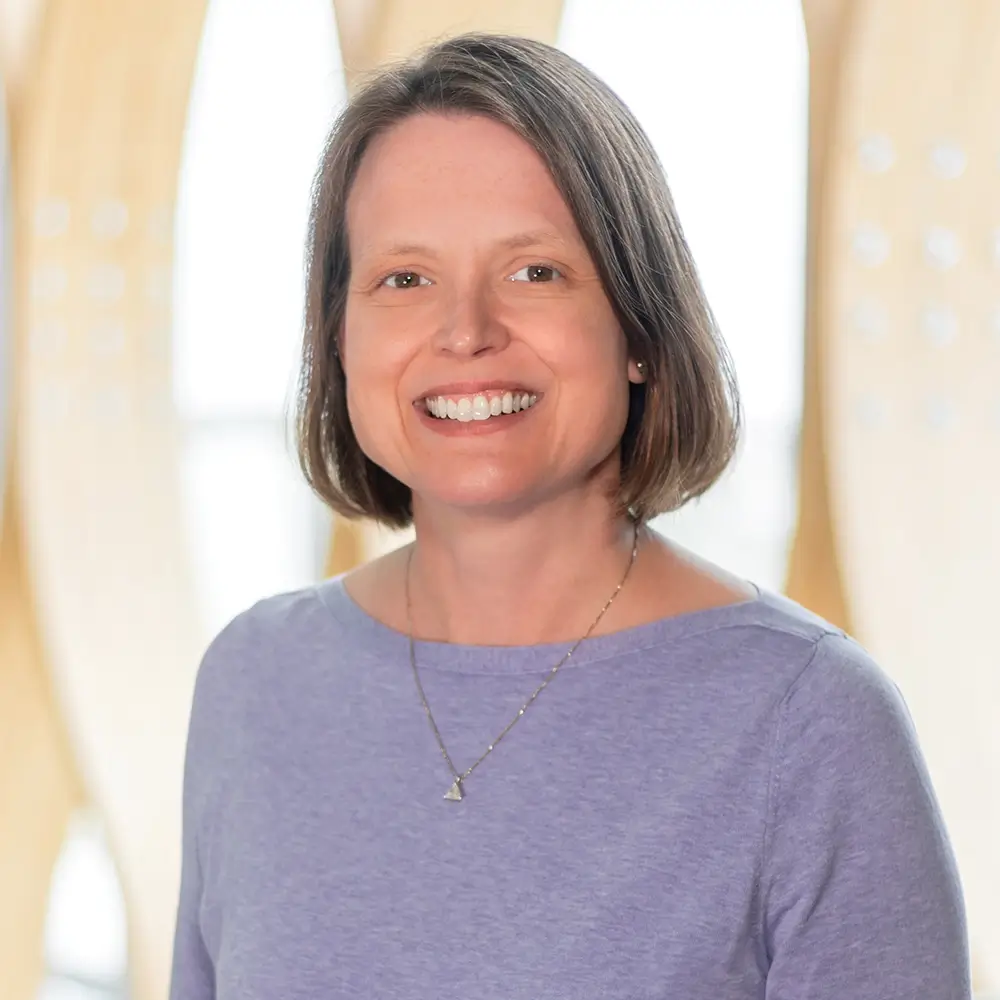 Dr. Krista Hill, MD, with short brown hair, wearing a lavender top and a delicate necklace, smiles at the camera while standing in front of a softly blurred light-colored background.