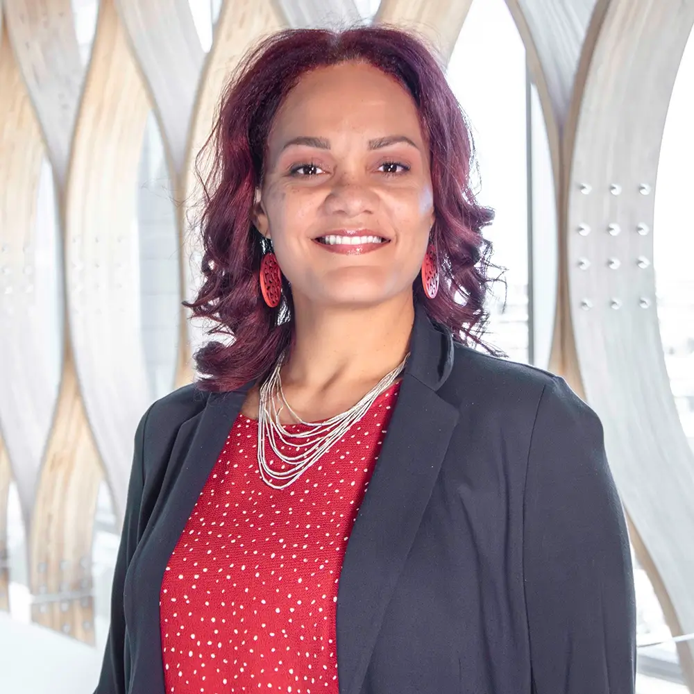 Dr. Lucille Woodard, MD, with shoulder-length curly hair, wearing a black blazer over a red polka-dot top, layered necklaces, and red earrings, stands smiling in front of a modern wooden architectural background.
