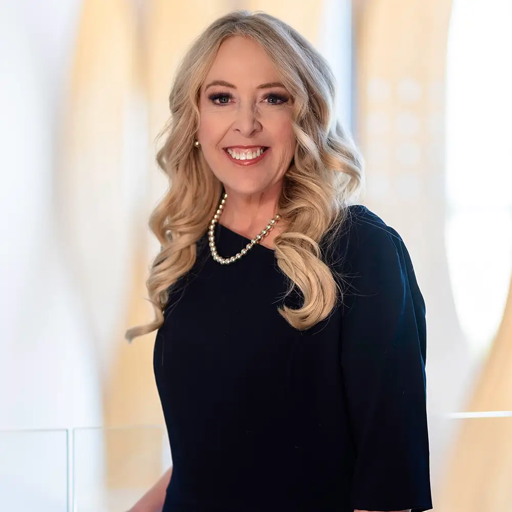 A smiling woman with long, wavy blonde hair, wearing a black dress and a pearl necklace, stands indoors against a light, blurred background—Margo Anderson-Fowler, MD.