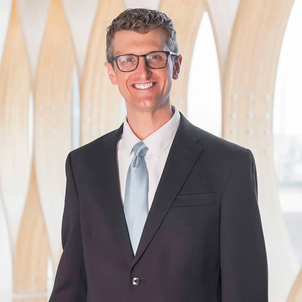 Dr. Mark Hare, MD, a smiling man wearing glasses, a dark suit, white shirt, and light blue tie, stands in front of a modern, light wooden architectural background.