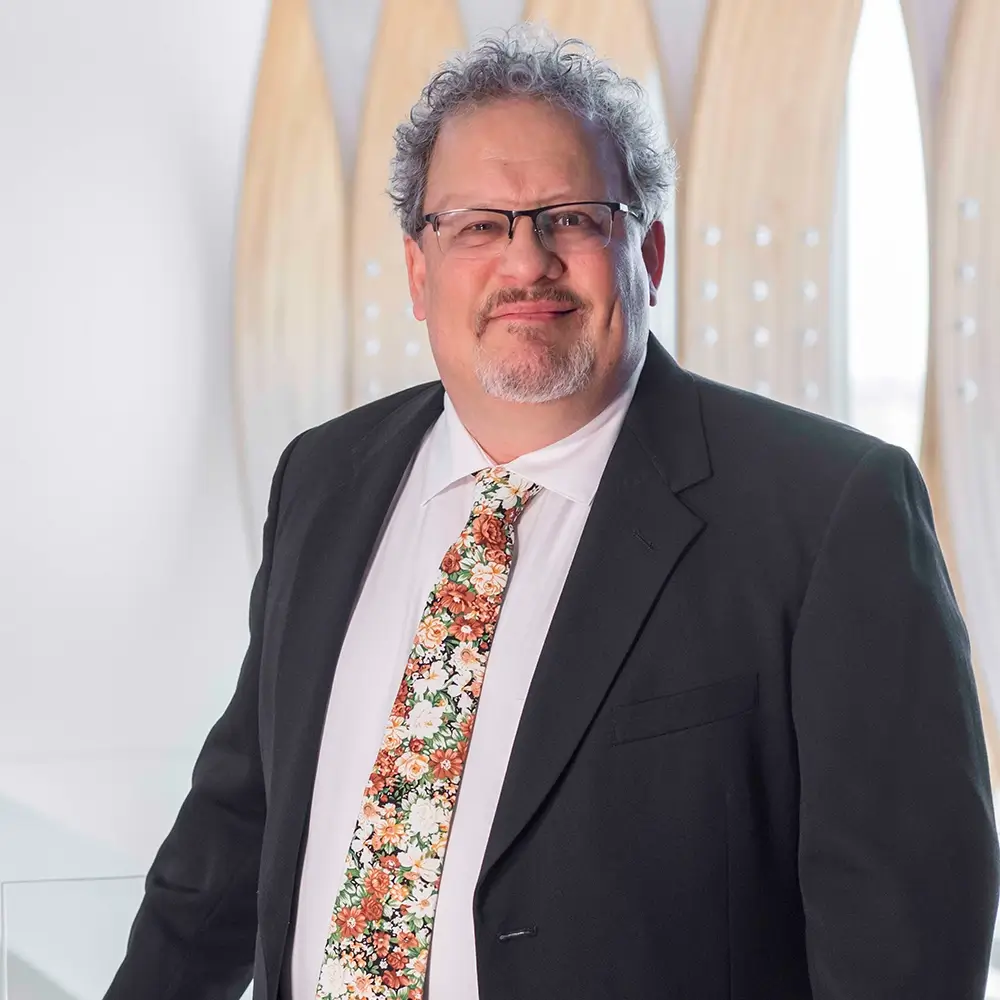 Mark Woodruff, MD, a man with curly gray hair, glasses, and a goatee, wears a black suit, white shirt, and a colorful floral tie while standing indoors in front of a light-colored, modern background.