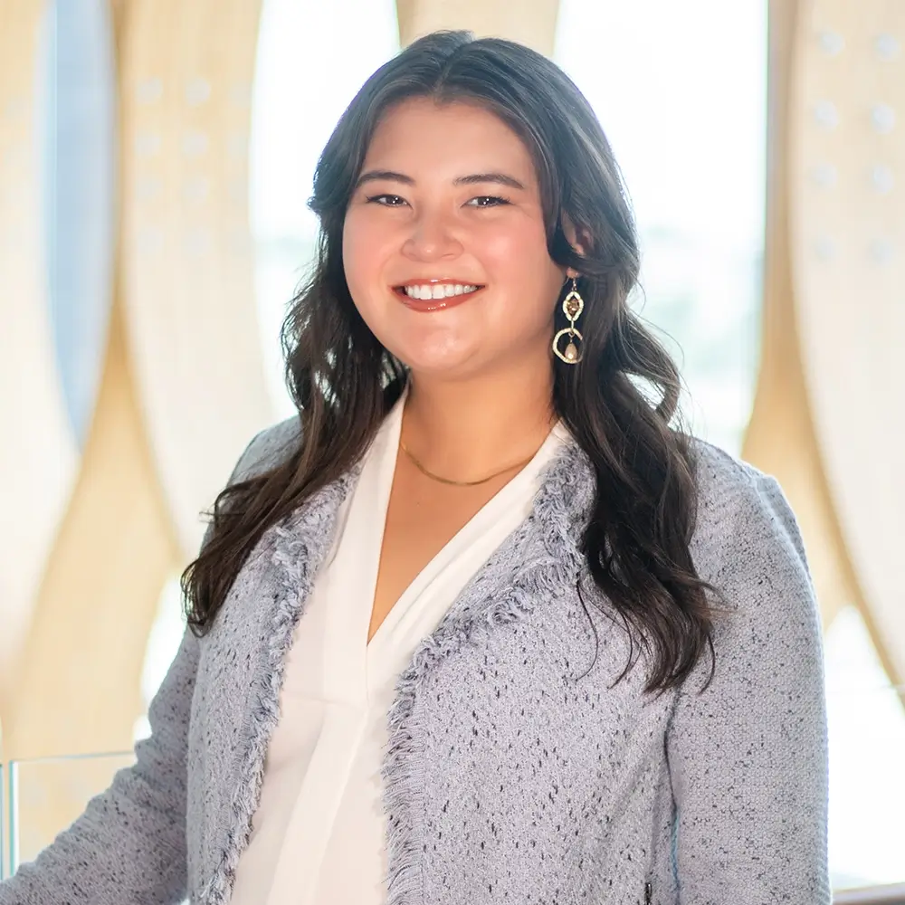 Melanie Kim, DO, with long dark hair, wearing a light gray textured blazer, white blouse, and gold earrings, stands smiling in front of a softly lit background with pale vertical patterns.