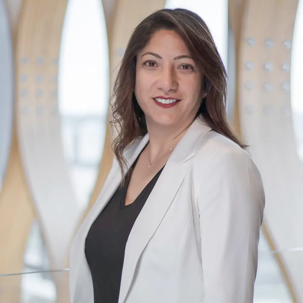 Dr. Nada Skaf, MD, with long brown hair, stands smiling in a white blazer over a black top against a modern, light-colored backdrop featuring curved wooden structures.