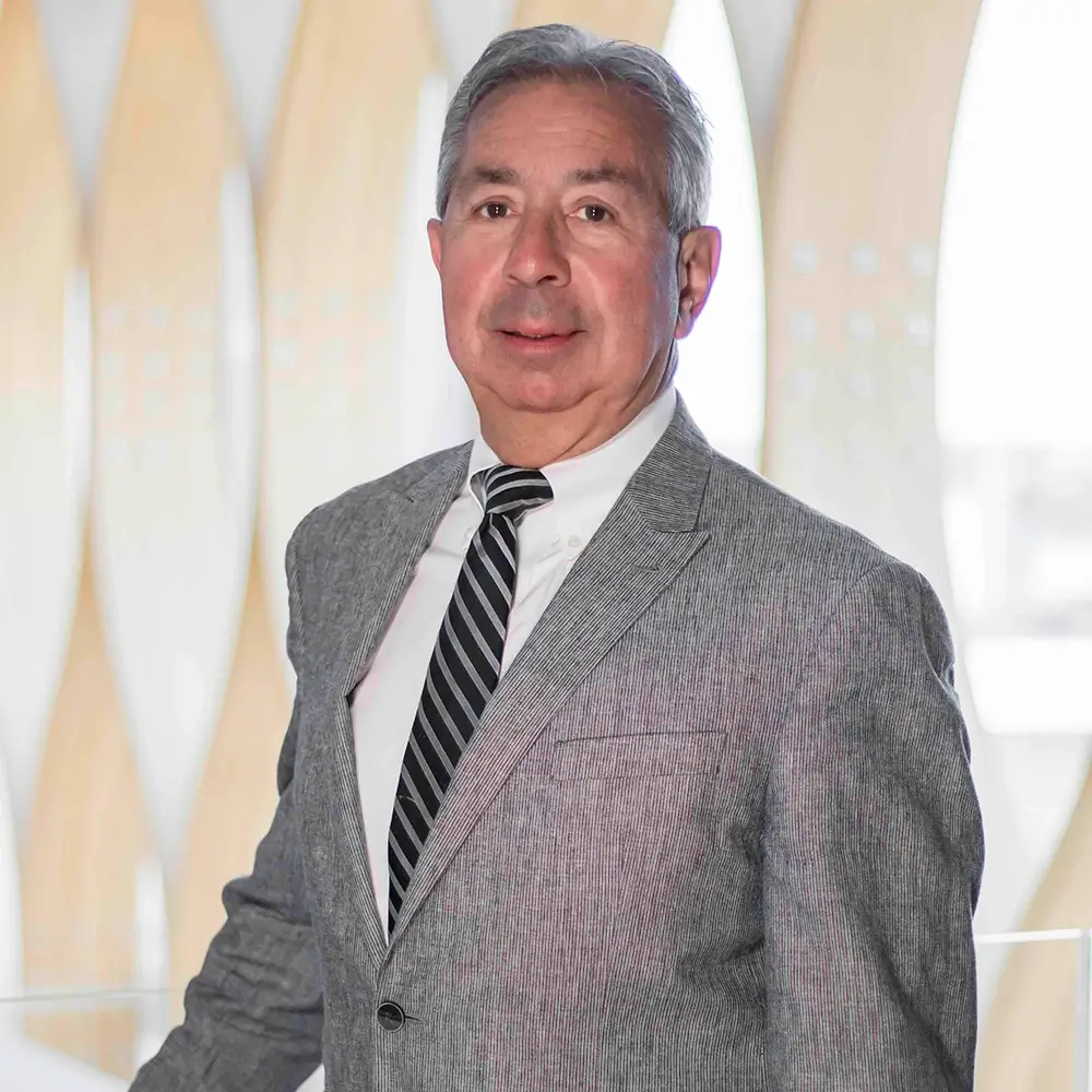 Steven Gradowski, a middle-aged man in a light gray suit, white shirt, and striped tie, stands indoors before a modern, softly-lit background with curved wooden panels. He faces the camera and smiles slightly, embodying professionalism in OD.